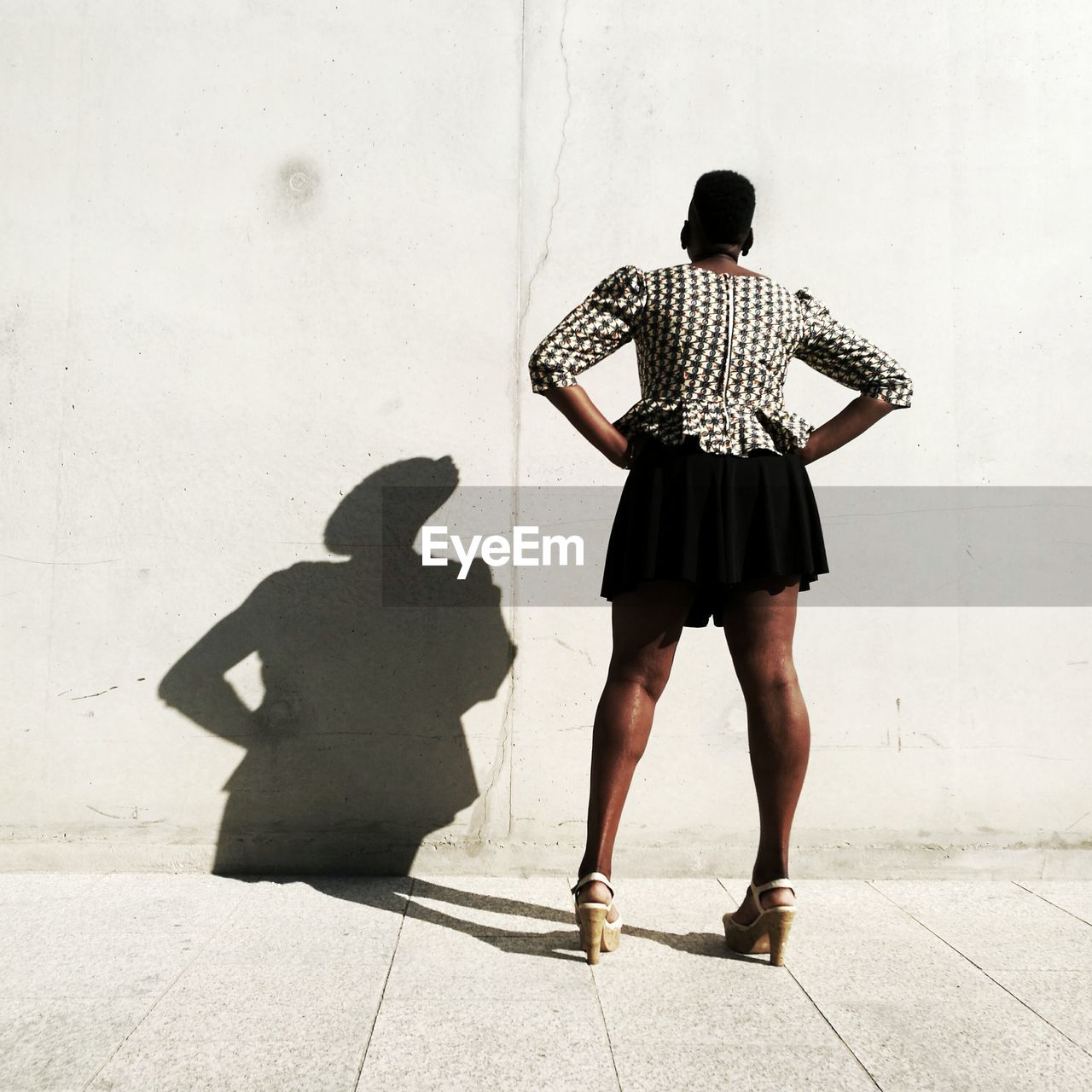 Rear view of woman with hands on hip standing against wall on sunny day