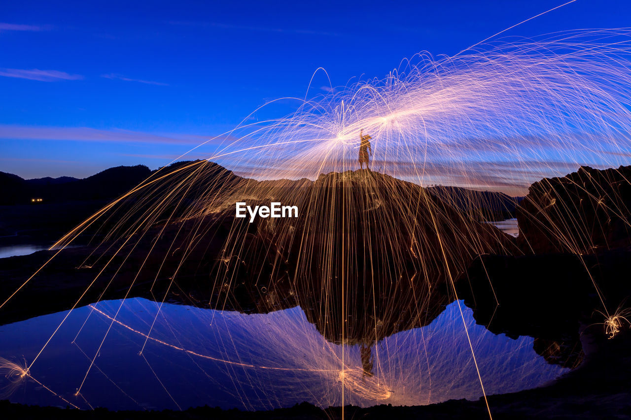 Silhouette man spinning wire wool on rock against sky at dusk