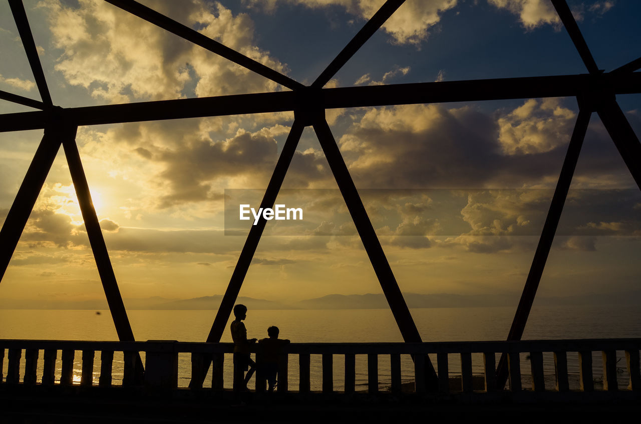 Silhouette boys by sea on bridge during sunset