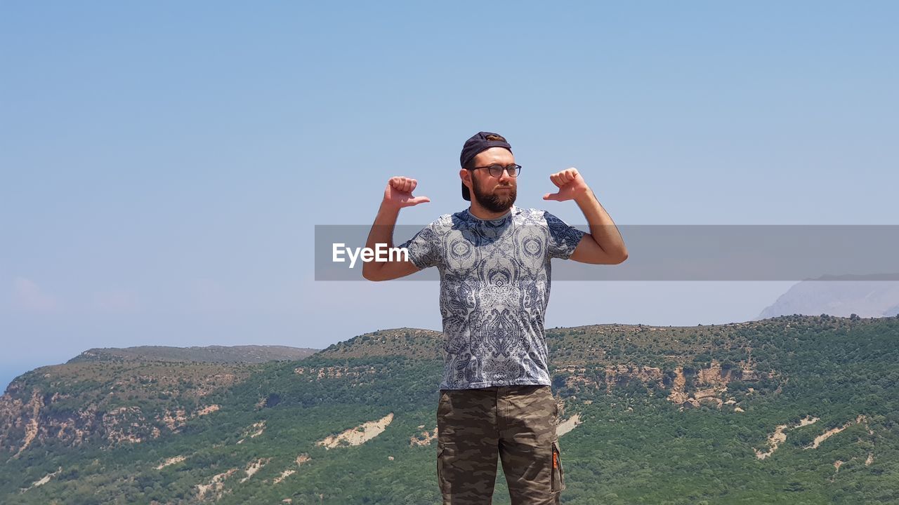 Young man gesturing thumbs up while standing on mountain against clear sky during sunny day