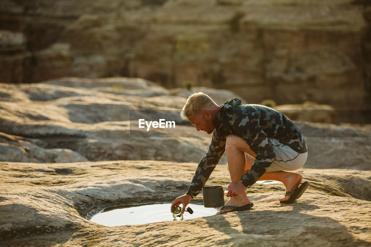 Man in camo jacket collects cooking water from a shallow puddle