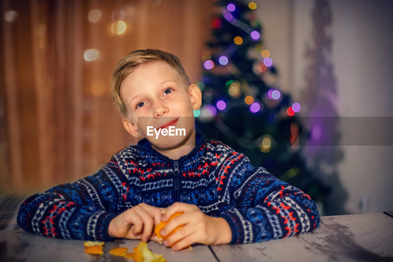 Portrait of boy holding orange sitting against christmas tree