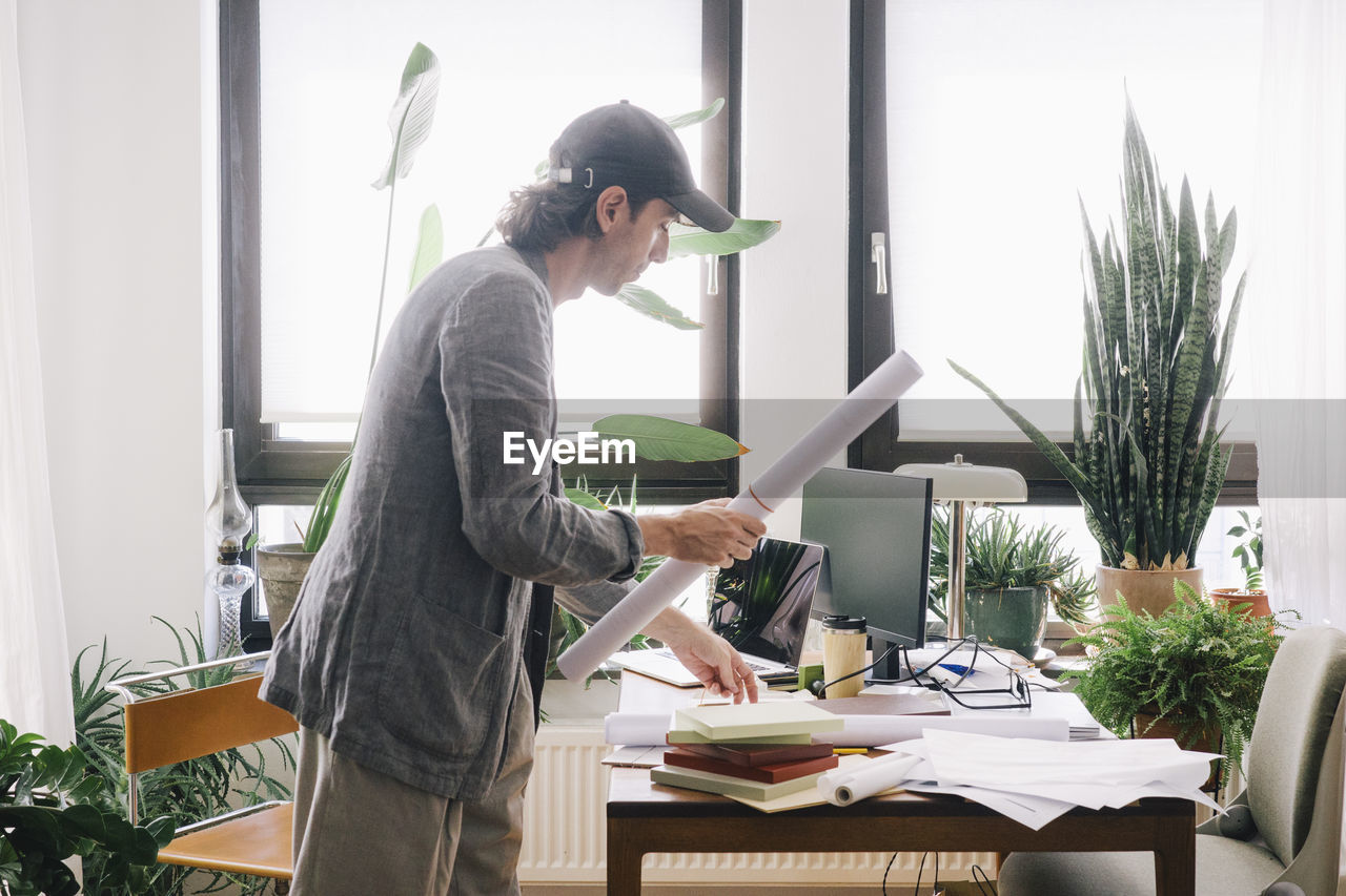 Side view of male architect holding rolled blueprint while standing at desk in home office