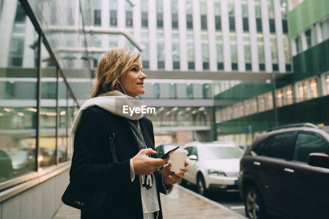 Businesswoman with coffee and smart phone looking away while standing on sidewalk against modern building in city