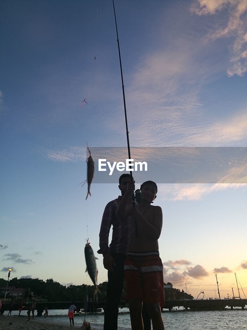 Silhouette people holding fish in rod while standing against sky during sunset