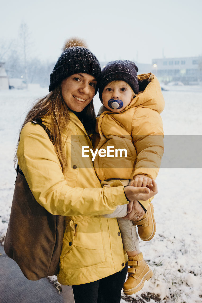 Portrait of smiling mother carrying son o footpath during winter