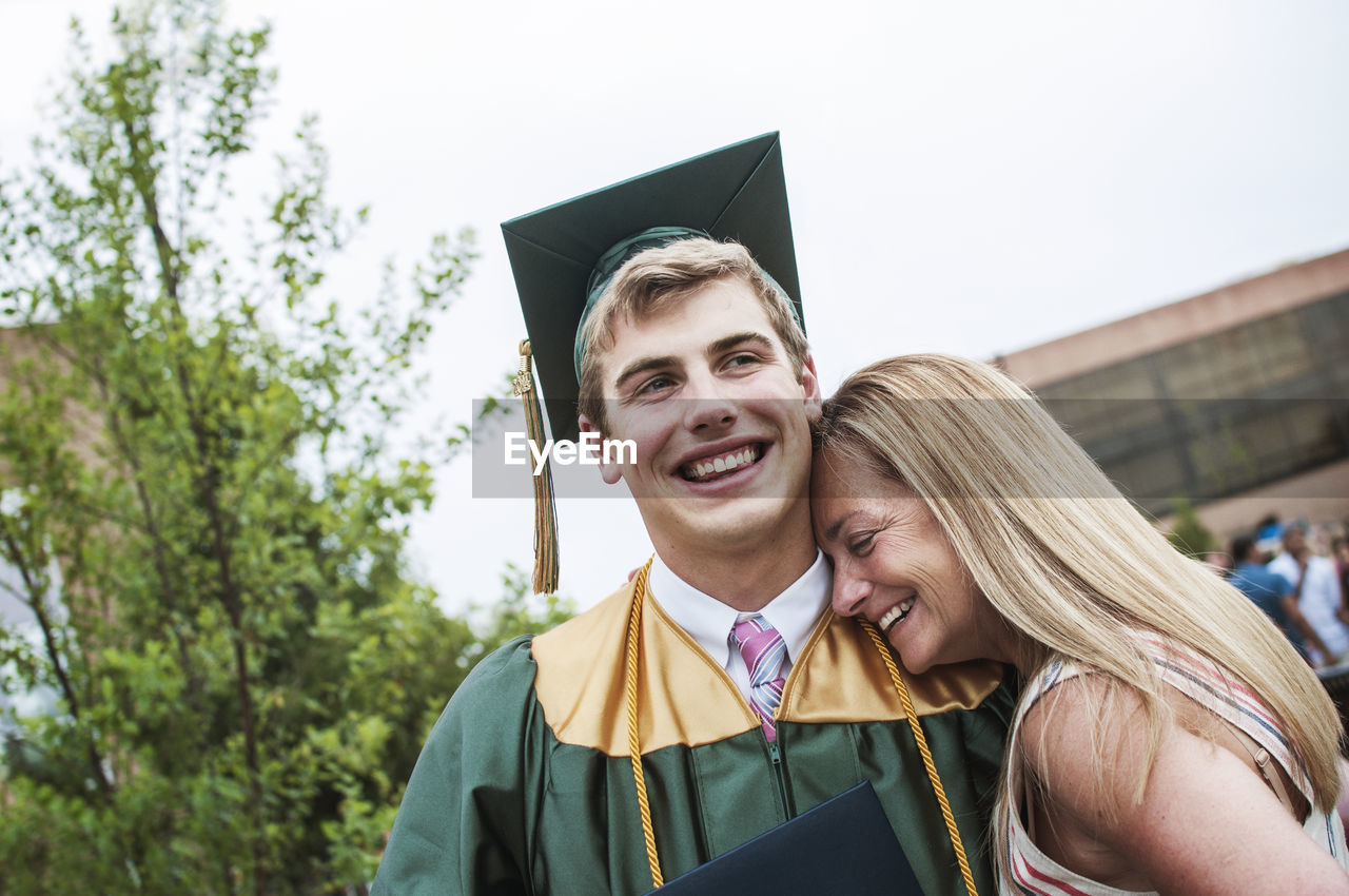 Happy mother leaning head on sons shoulder at graduation ceremony