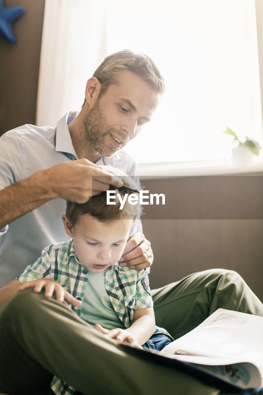 Boy reading book while father combing his hair at home