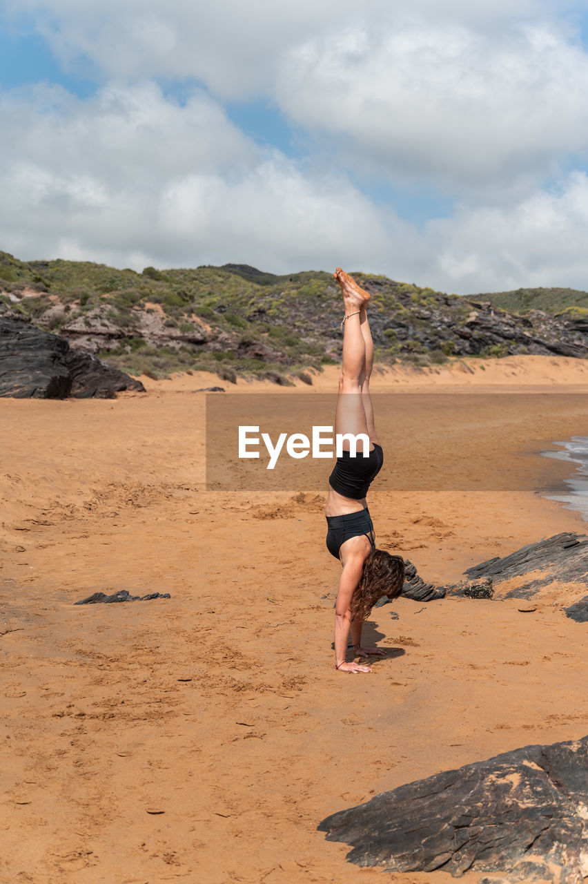 Side view of woman doing adho mukha vriksasana on stone against grassy hills and cloudy sky during yoga session on sandy beach in summer