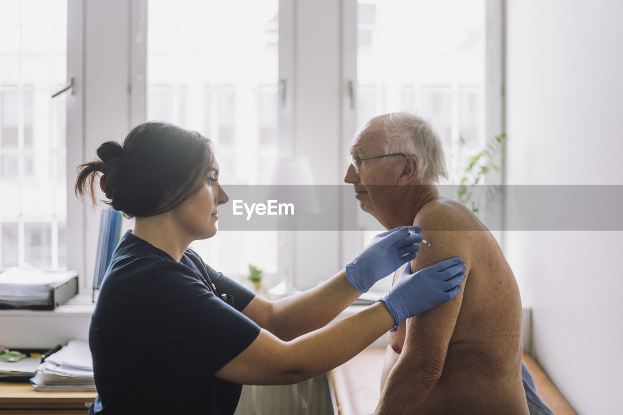 Female nurse giving vaccination to shirtless senior patient at hospital