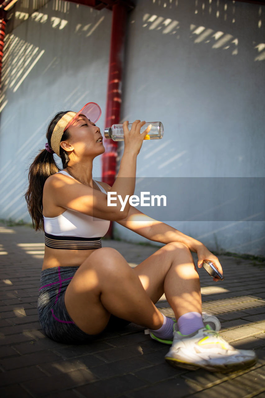 Portrait of young asian woman sitting on floor after training and drinking water