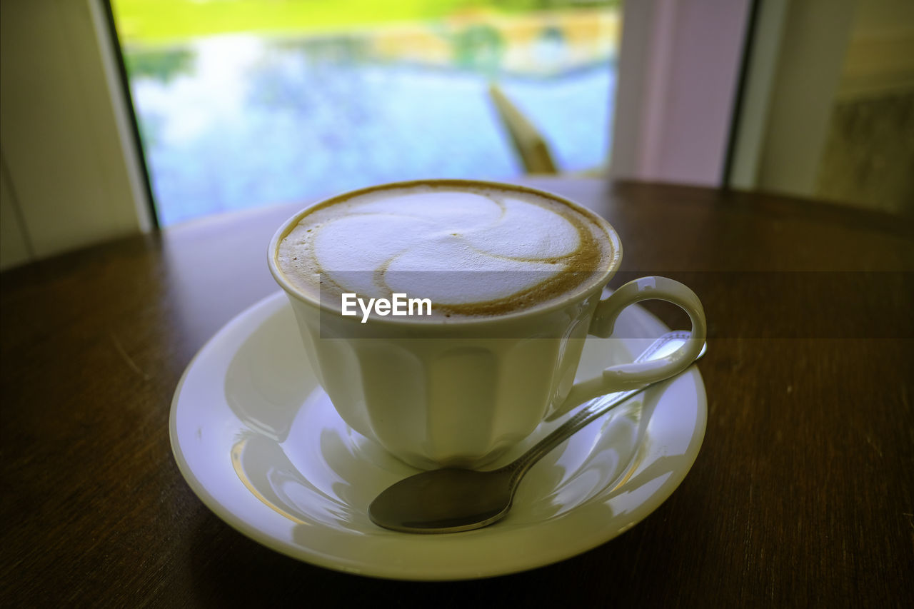 A white cup of hot coffee with floral latte art, serving on brown table in natural light near window