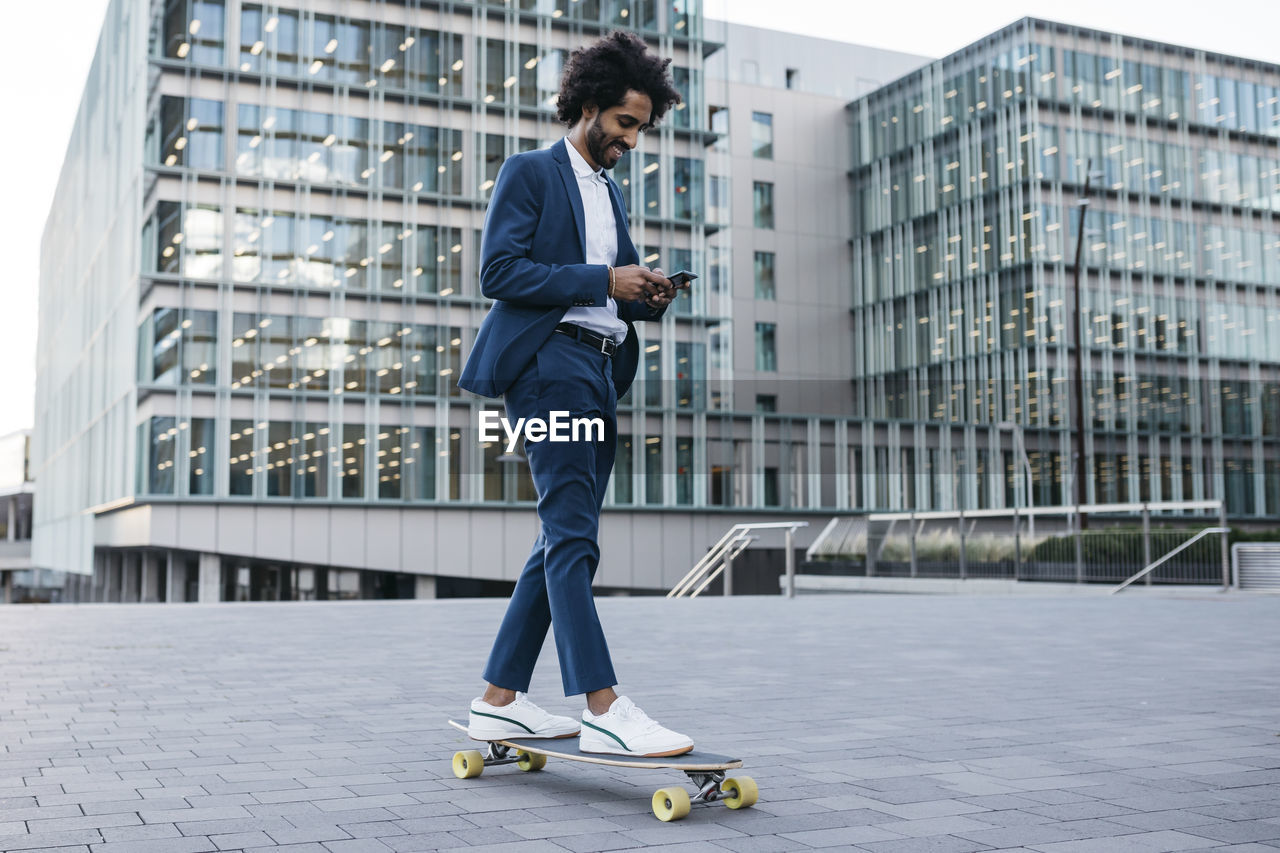 Spain, barcelona, young businessman riding skateboard and using cell phone in the city