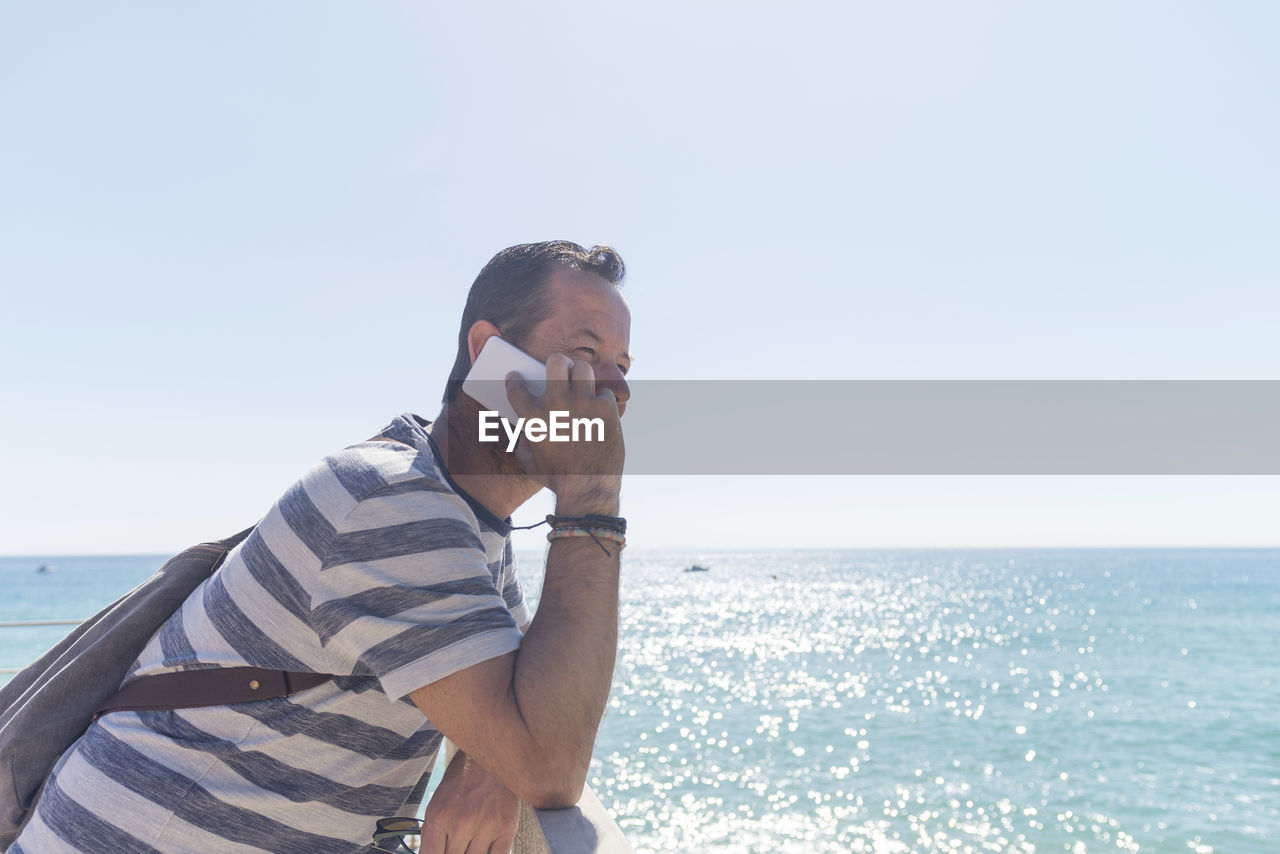 Side view of man listening over phone while standing at beach against clear sky