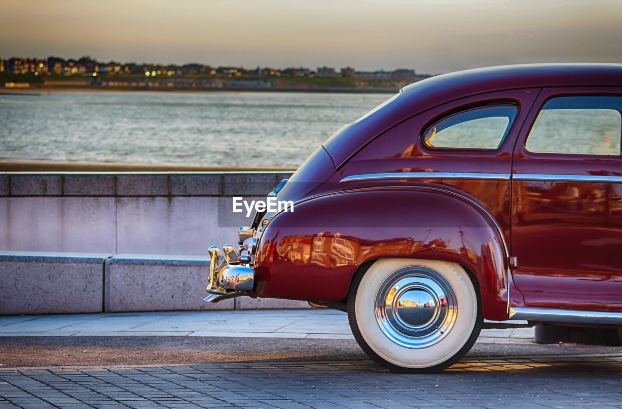 Cropped image of vintage car on street by river against sky during sunset