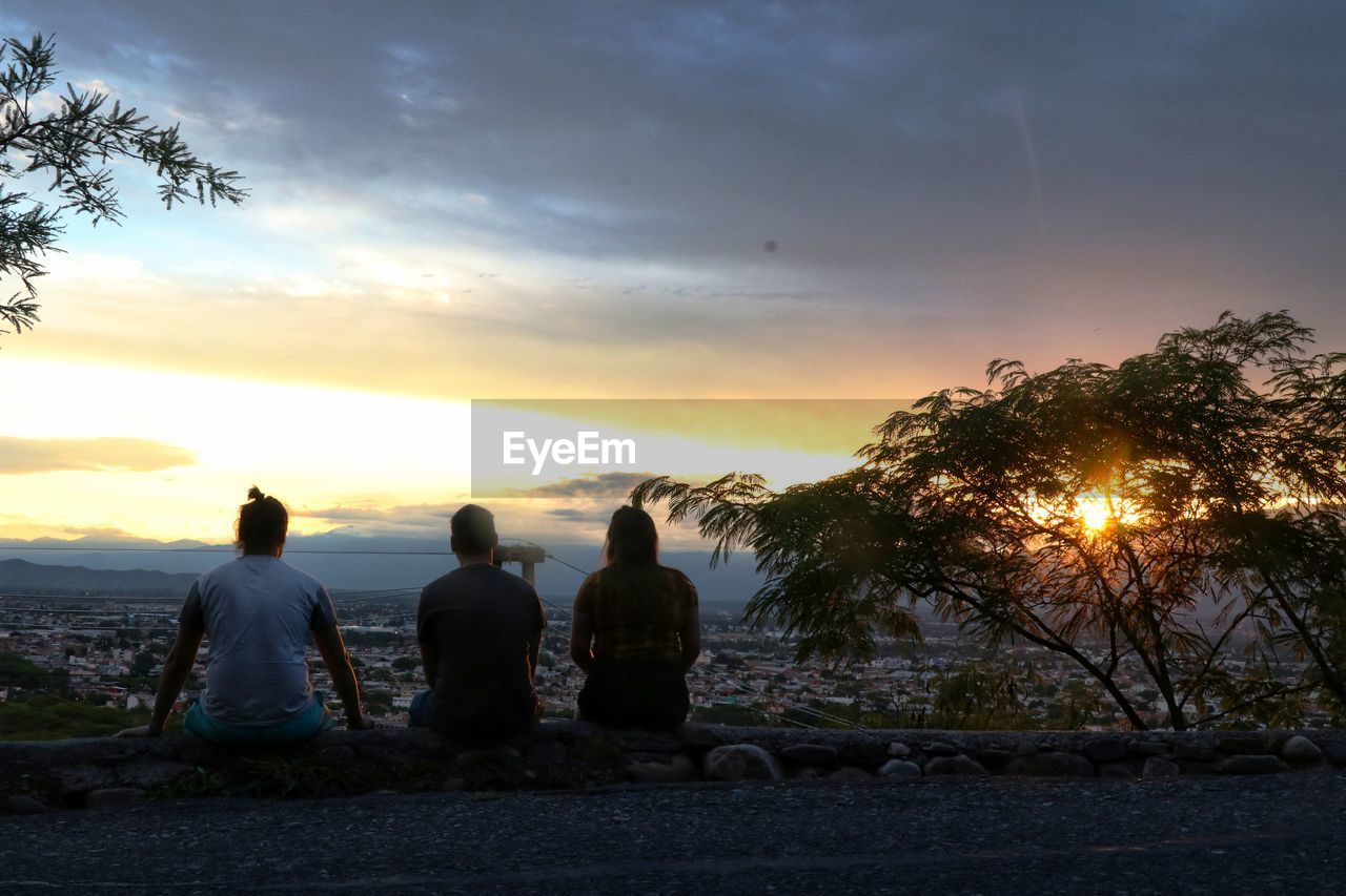 REAR VIEW OF PEOPLE SITTING ON SHORE AGAINST SKY