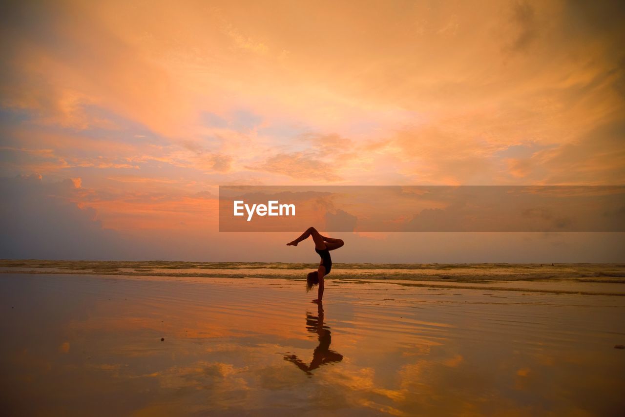 Silhouette teenage girl doing handstand at beach against cloudy sky during sunset