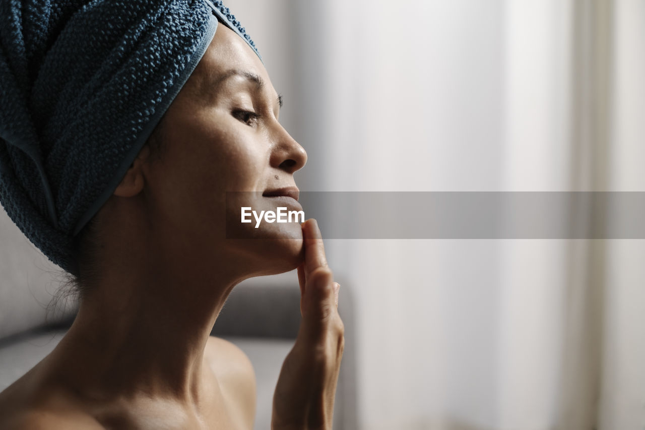 Side view of charming young female with bare shoulders and towel on head smearing facial skincare cream on face during makeup procedure at home