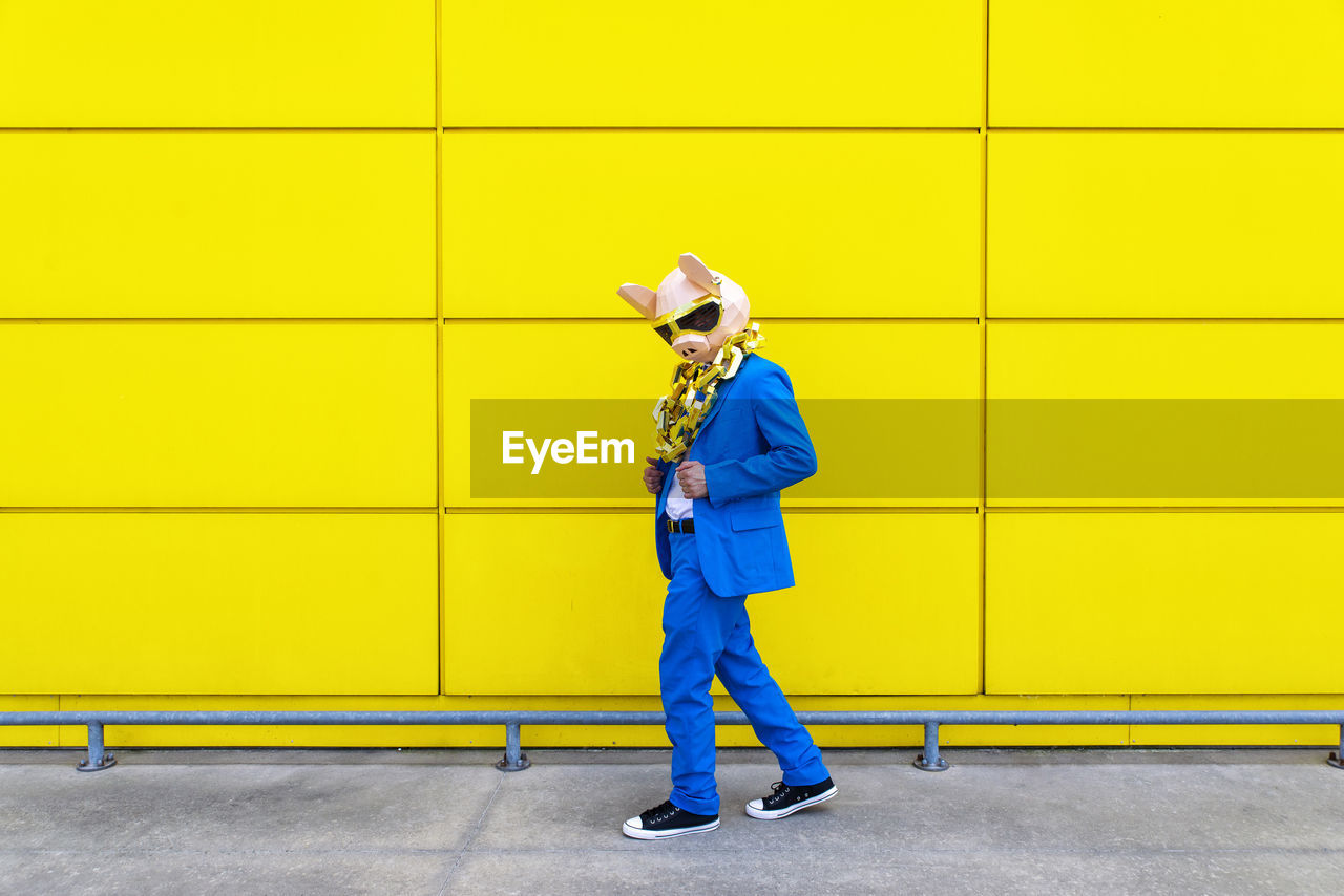 Man wearing vibrant blue suit, pig mask and large golden chain posing against yellow wall