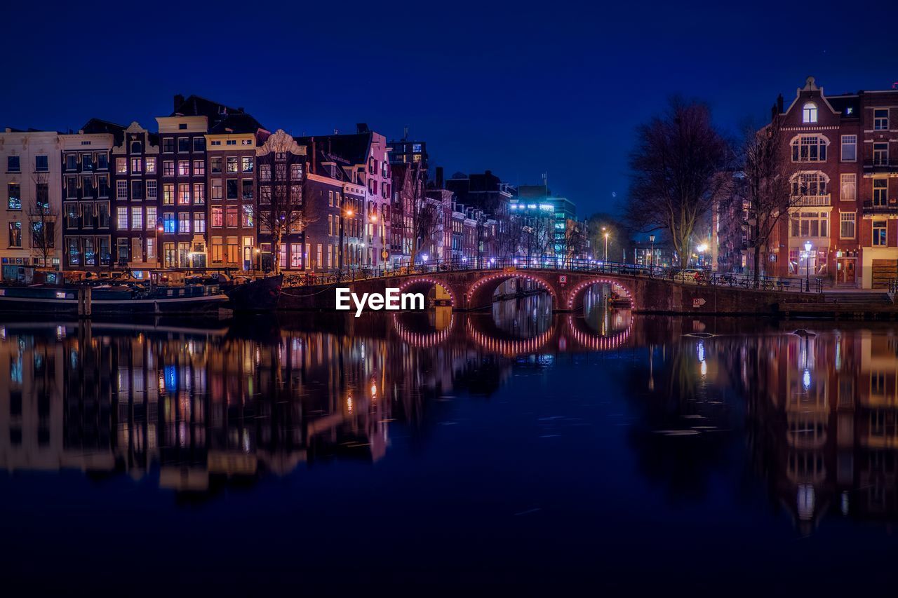 Illuminated bridge over river by buildings against sky at night