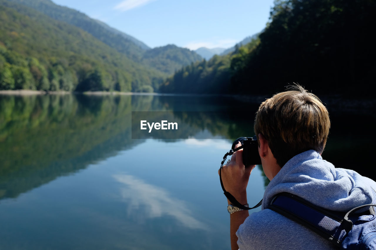 Rear view of woman photographing by lake against sky