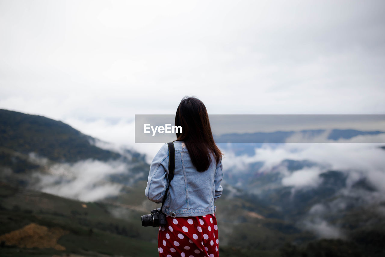 Rear view of woman with camera standing on mountain against sky