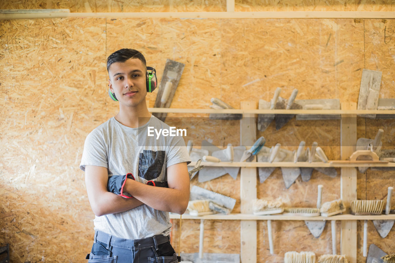 Portrait of confident teenage trainee standing with arms crossed against wall at workshop