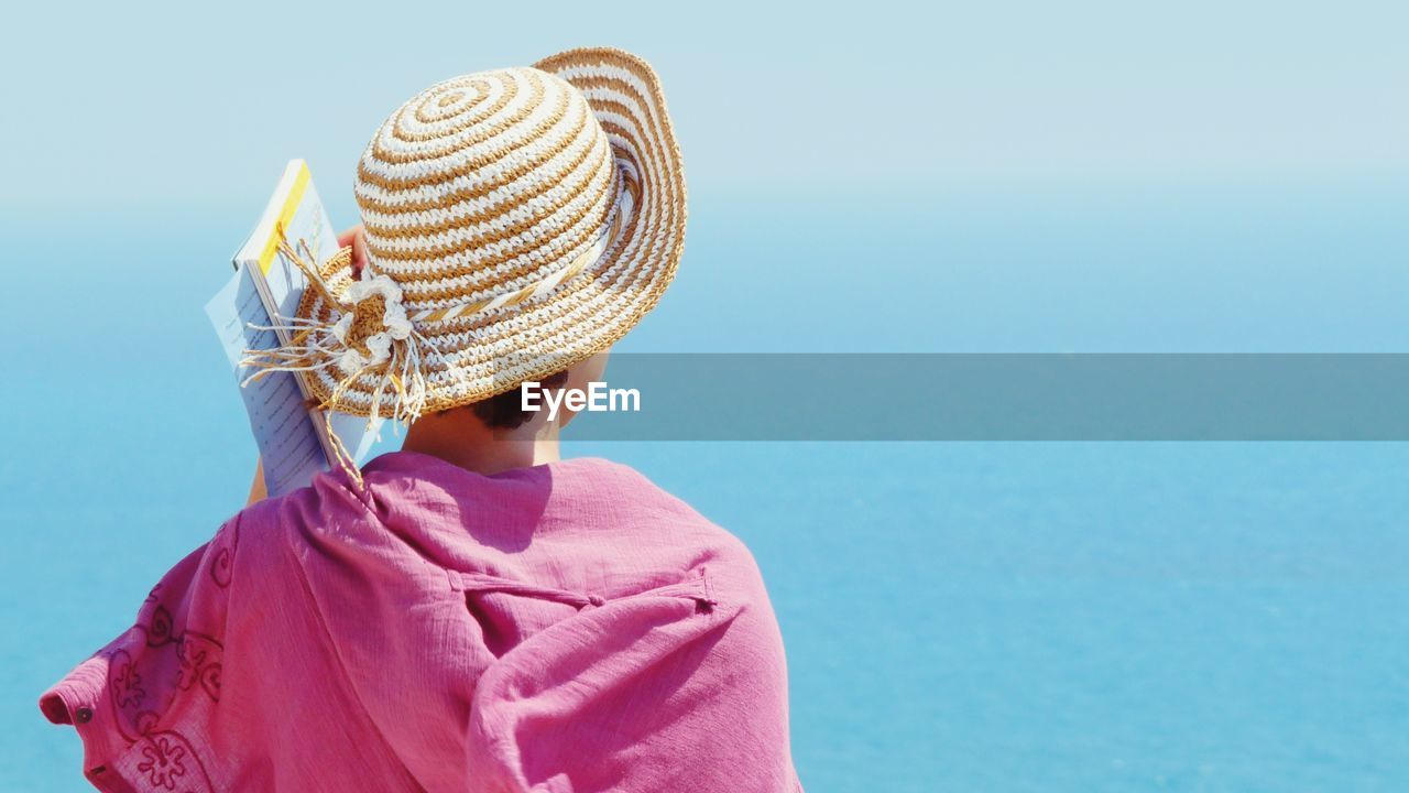 Rear view of woman with books wearing hat against clear blue sky