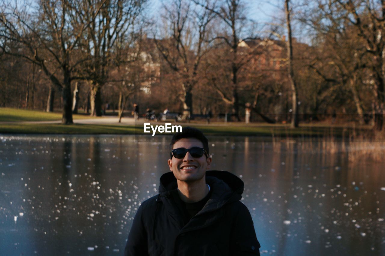 Portrait of smiling young man wearing sunglasses standing against lake