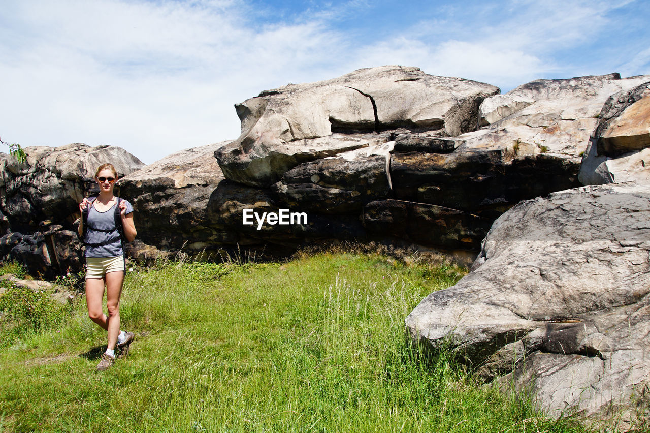 Female hiker walking on grassy field by rock formation against sky during sunny day