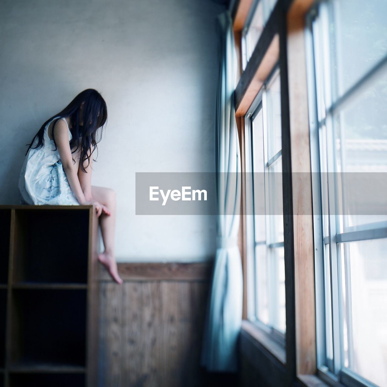 Side view of woman sitting on cabinet against wall at home