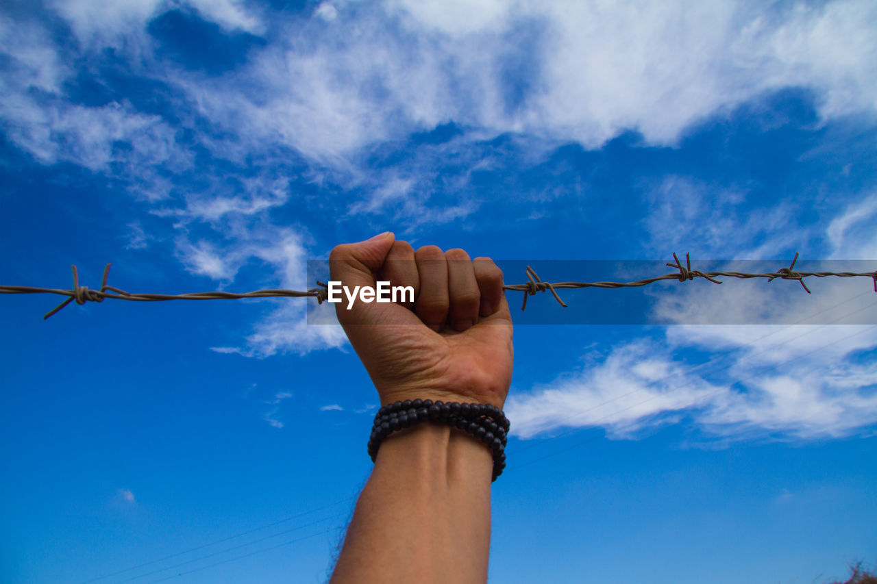 Cropped hand of man holding barbed wire against blue sky