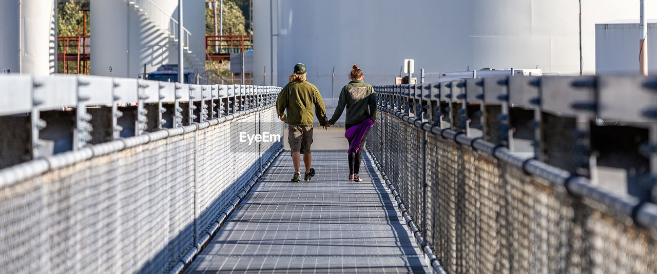 REAR VIEW OF PEOPLE WALKING ON BRIDGE AGAINST BUILDINGS