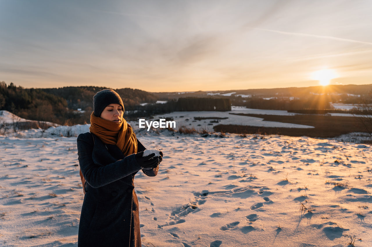 Young woman in coat and hat at sunset stands on snow-covered field, holds handful of snow in hands