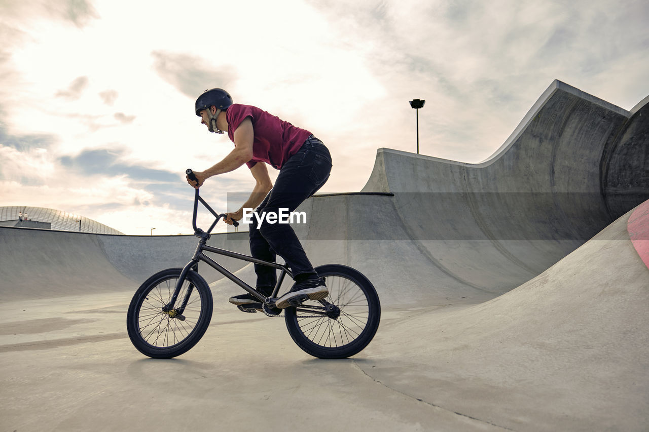 Rider riding bmx bike on concrete ramp against cloudy sky at skateboard park