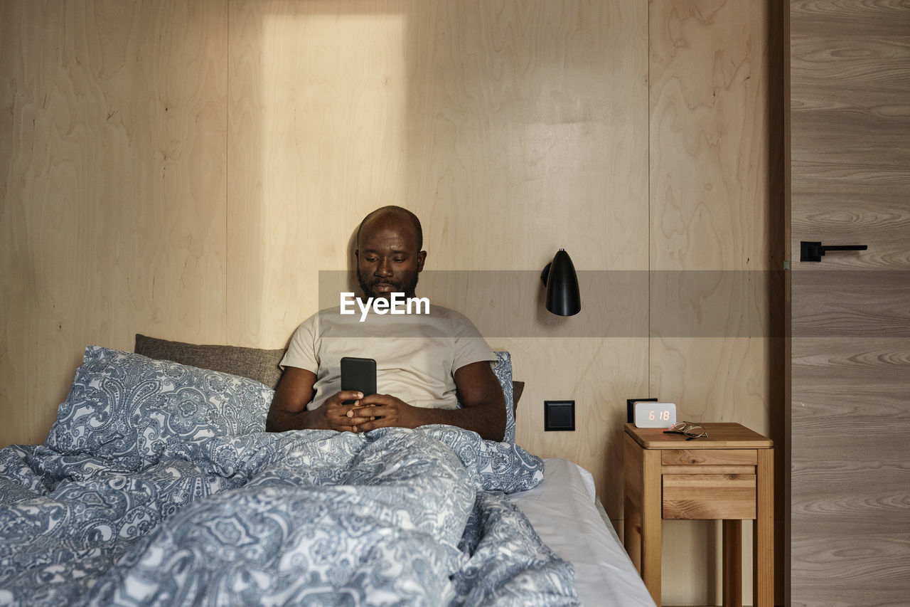 Young man using smart phone sitting on bed in bedroom at home