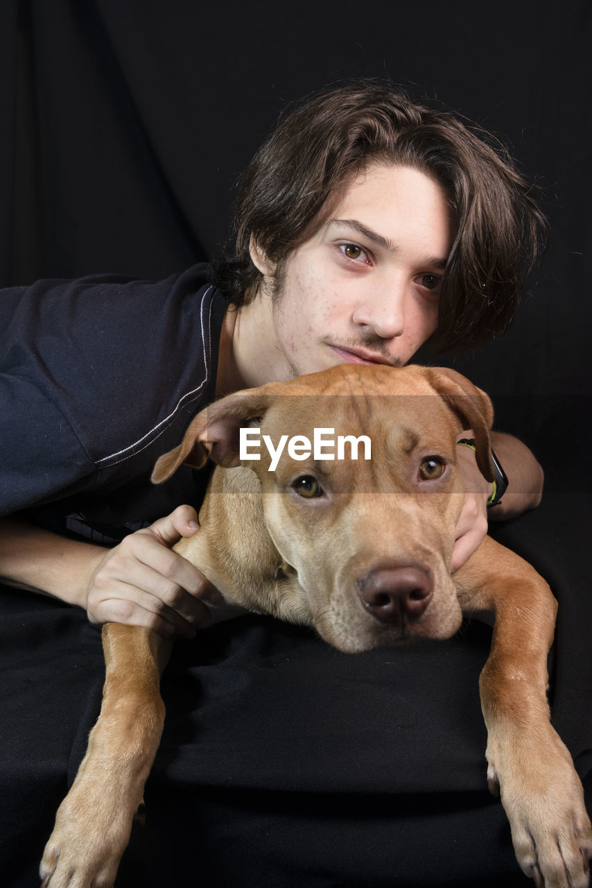Portrait of a young man with a caramel colored pitbull dog against black background. 