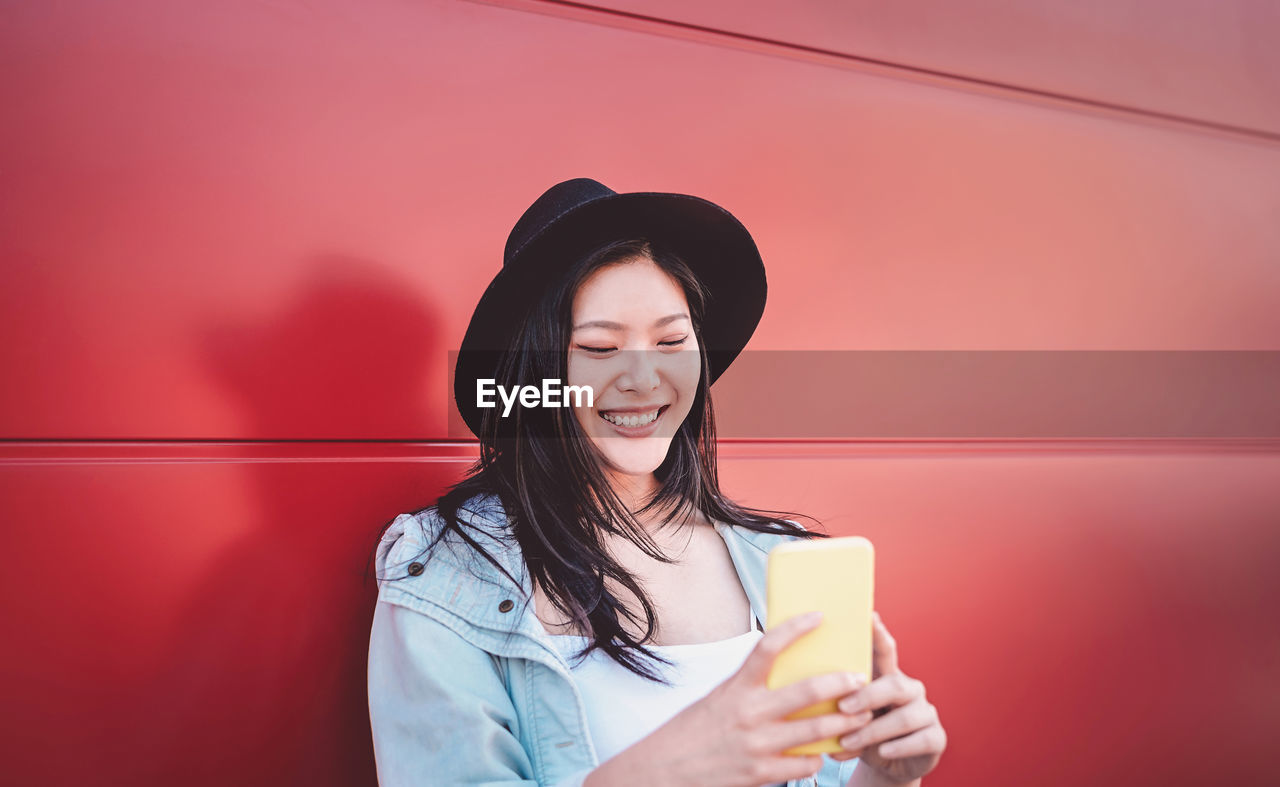 Smiling young woman using mobile phone against wall