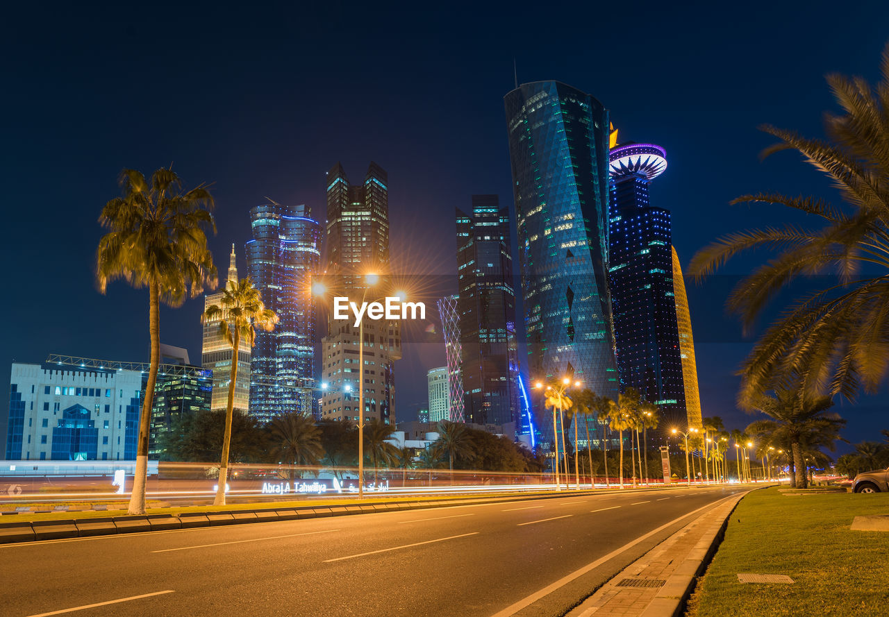 LIGHT TRAILS ON ROAD BY BUILDINGS AGAINST SKY AT NIGHT