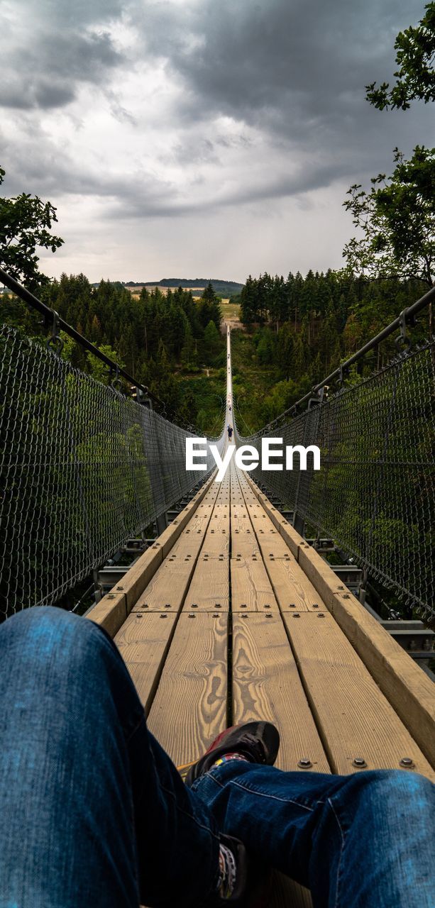 Low section of man relaxing on footbridge against cloudy sky