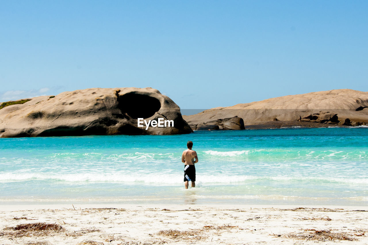 water, sea, land, beach, vacation, shore, ocean, coast, sky, one person, body of water, sand, nature, wave, full length, beauty in nature, holiday, trip, leisure activity, day, scenics - nature, clear sky, sunny, blue, bay, standing, adult, lifestyles, rock, rear view, men, outdoors, travel destinations, travel, sunlight, tranquility, idyllic, tranquil scene, walking, motion, person, wind wave, non-urban scene, women