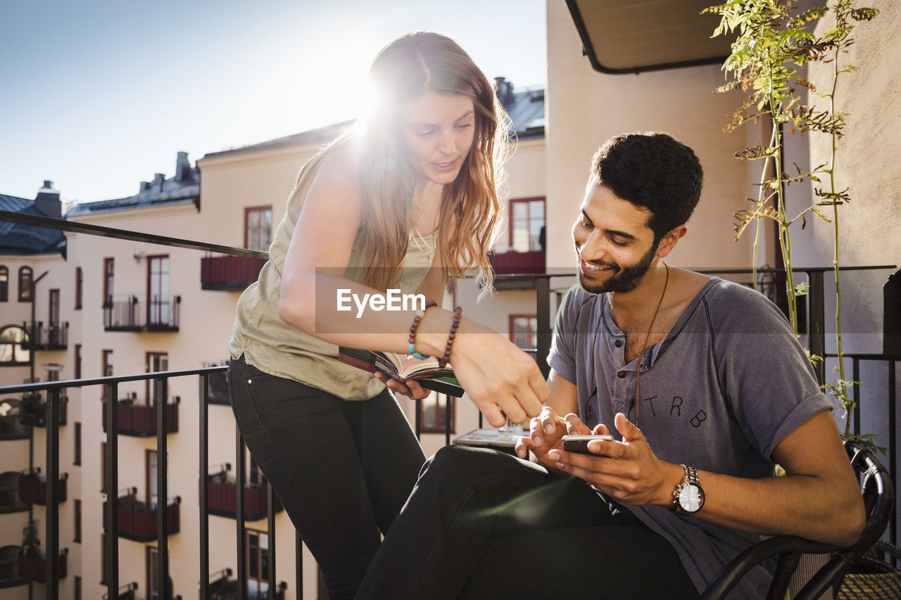 Happy couple using smart phone while reading guidebook on balcony