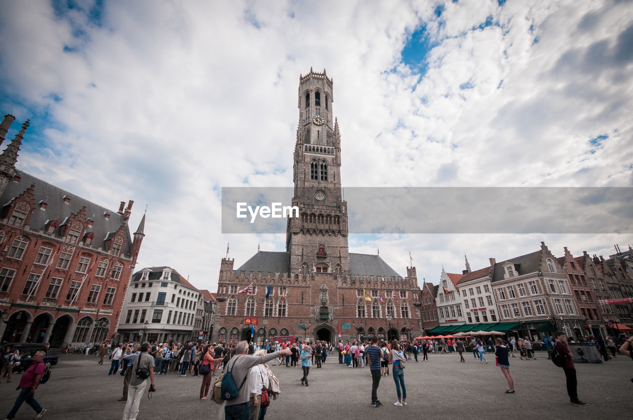 People in front of medieval clock tower against cloudy sky