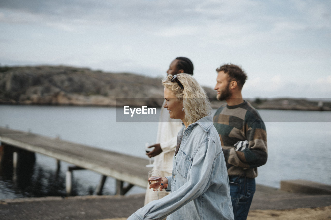 Side view of young woman enjoying drinks while walking with friends near lake