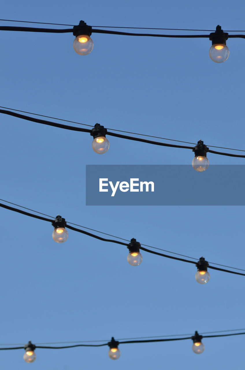 Strings of illuminated electric light bulbs outdoors against blue sky