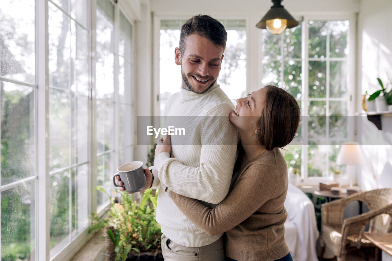 Side view of cheerful female embracing bearded male beloved with mug of coffee while looking away against window in house