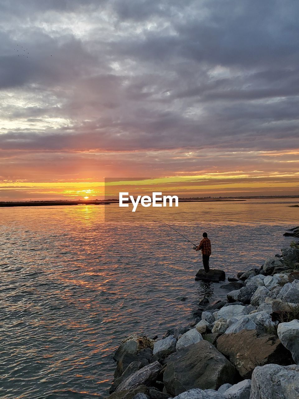 MAN STANDING ON ROCKS AT SHORE AGAINST SKY DURING SUNSET
