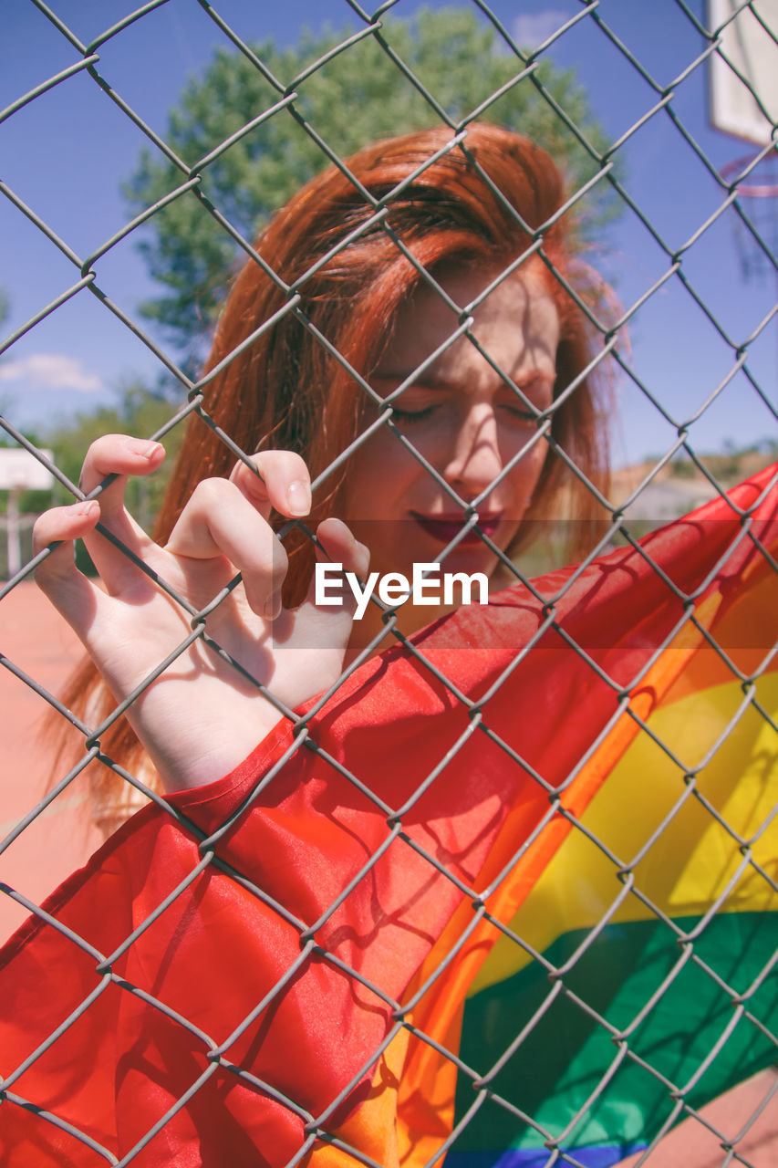 Young woman with rainbow flag seen through chainlink fence