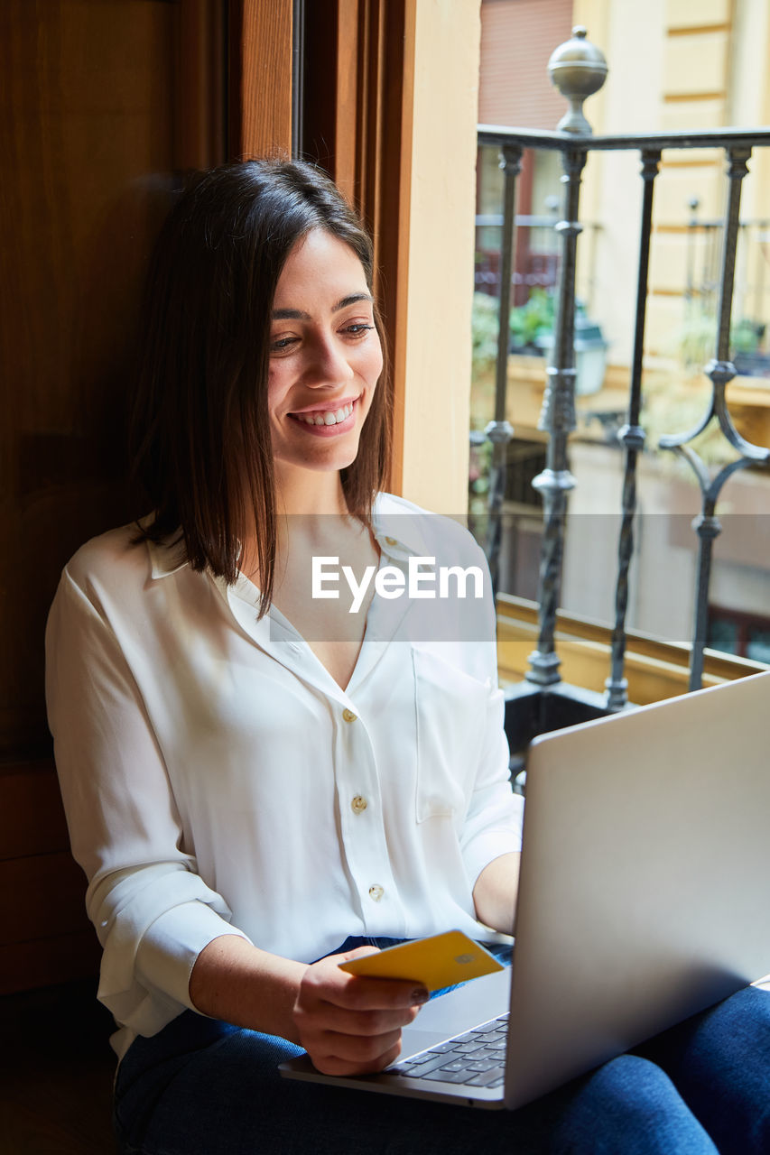 Young woman shopping and paying online on a laptop with credit card while relaxing at home next to a window.