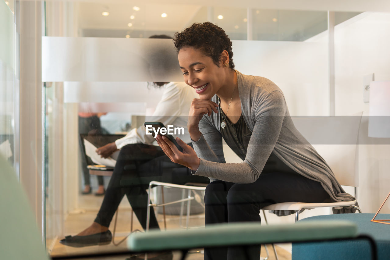 SMILING YOUNG WOMAN USING PHONE WHILE SITTING ON SEAT IN OFFICE